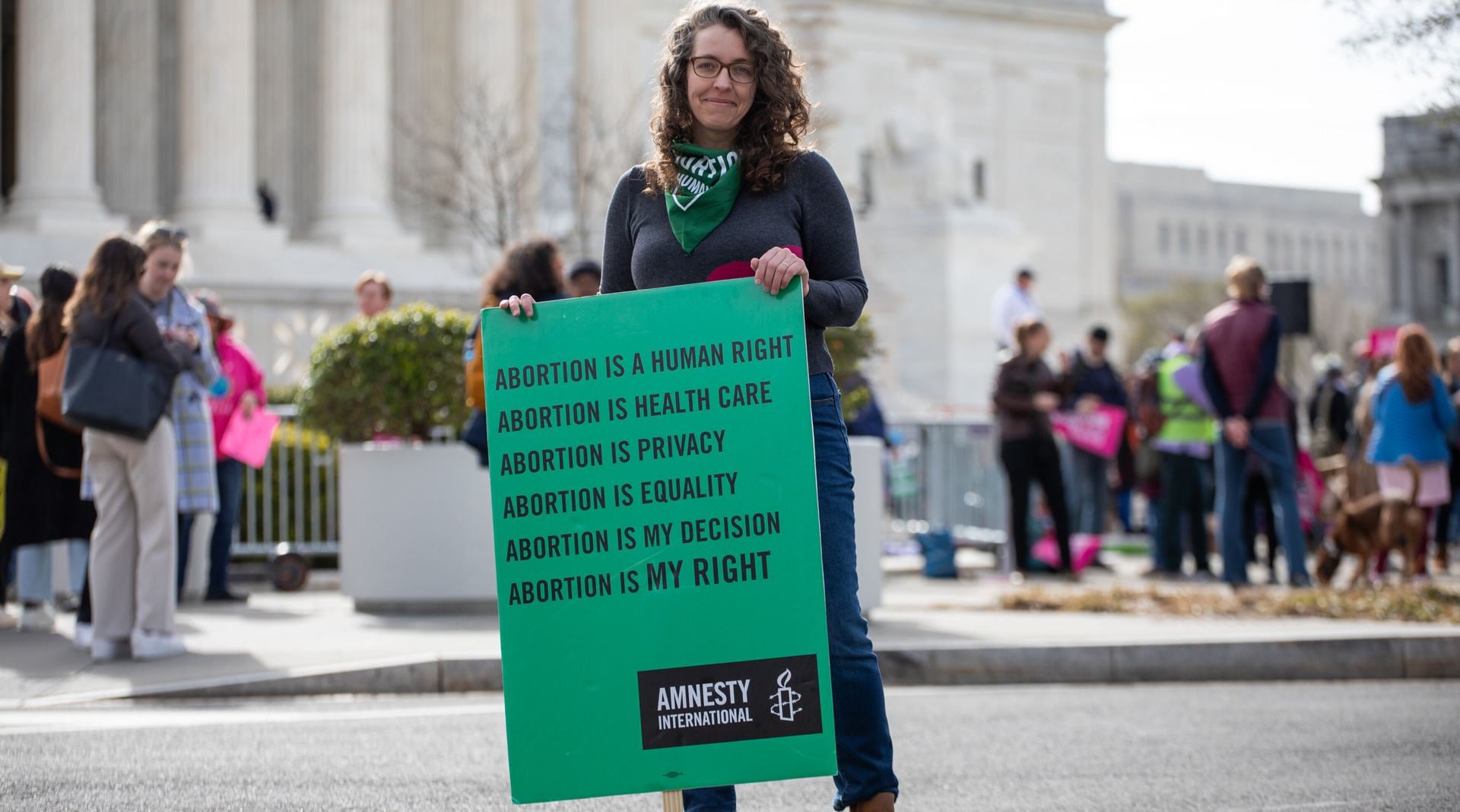 White woman stands outside a building with a green placard that reads "Abortion is a human right, abortion is health care, abortion is privacy, abortion is equality, abortion is my decision, abortion is MY RIGHT"