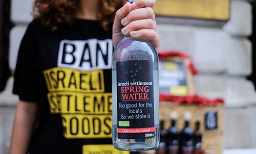 Person holding a bottle with the title "Israeli settlement Spring Water - Too good for the locals, so we stole it", and wearing a Ban Israeli Settlement Goods.