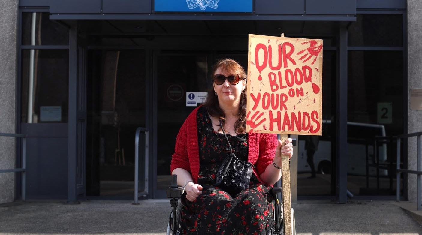 An activist outside of a government building, holding a banner that says "Our Blood, Your Hands"