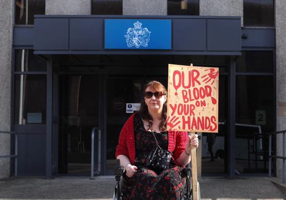 An activist outside of a government building, holding a banner that says "Our Blood, Your Hands"