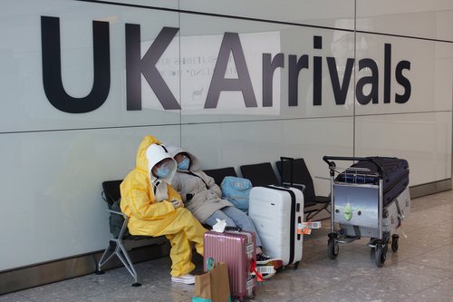 Two people in full PPE in a waiting area with their luggage with a 'UK Arrivals' sign behind them