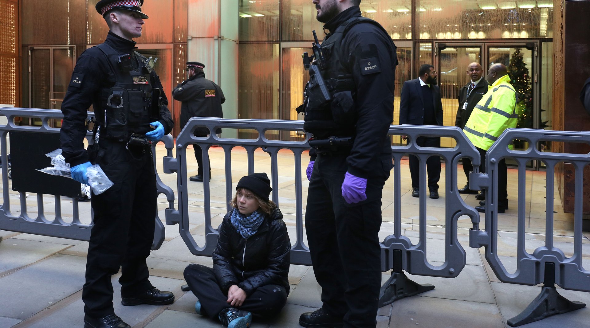 Greta Thunberg sits on a pavement with two male police officers standing either side of her in the process of arresting her.