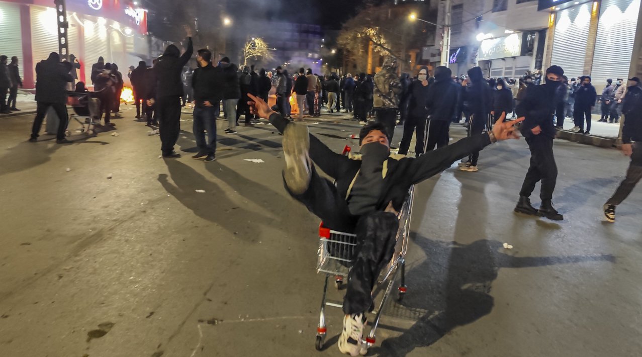 Hundreds of protesters on a street in Tehran at nigh time. One of the protesters has his arms raised in the air.