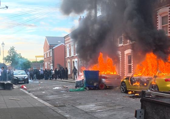 Flames and thick black smoke rise from a vehicle on a terraced street during racist riots in Belfast, Northern Ireland, June, 2026