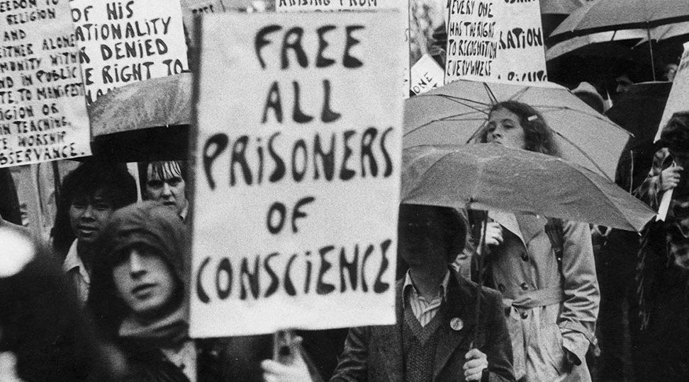 Black and white image of protesters from 1977 attending a demonstration holding a placard that says "Free all prisoners of conscience".