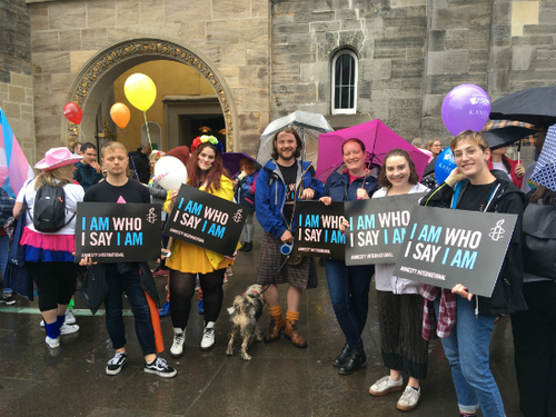 A group of six individuals from Amnesty International Scotland holding black signs with the text 'I Am Who I Say I Am' printed in pink and blue.