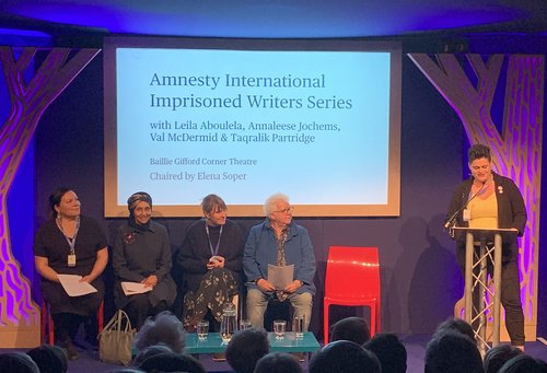 Image shows a woman reading at a lectern whilst four people sit on a stage