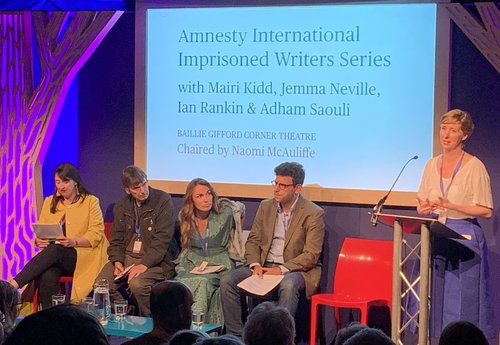 Image shows a woman reading at a lectern whilst four people sit on a stage