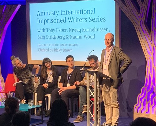 Image shows a man reading at a lectern whilst four people sit on a stage