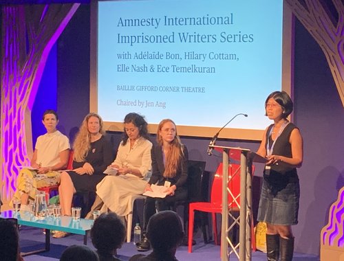 Image shows a woman reading at a lectern whilst four people sit on a stage