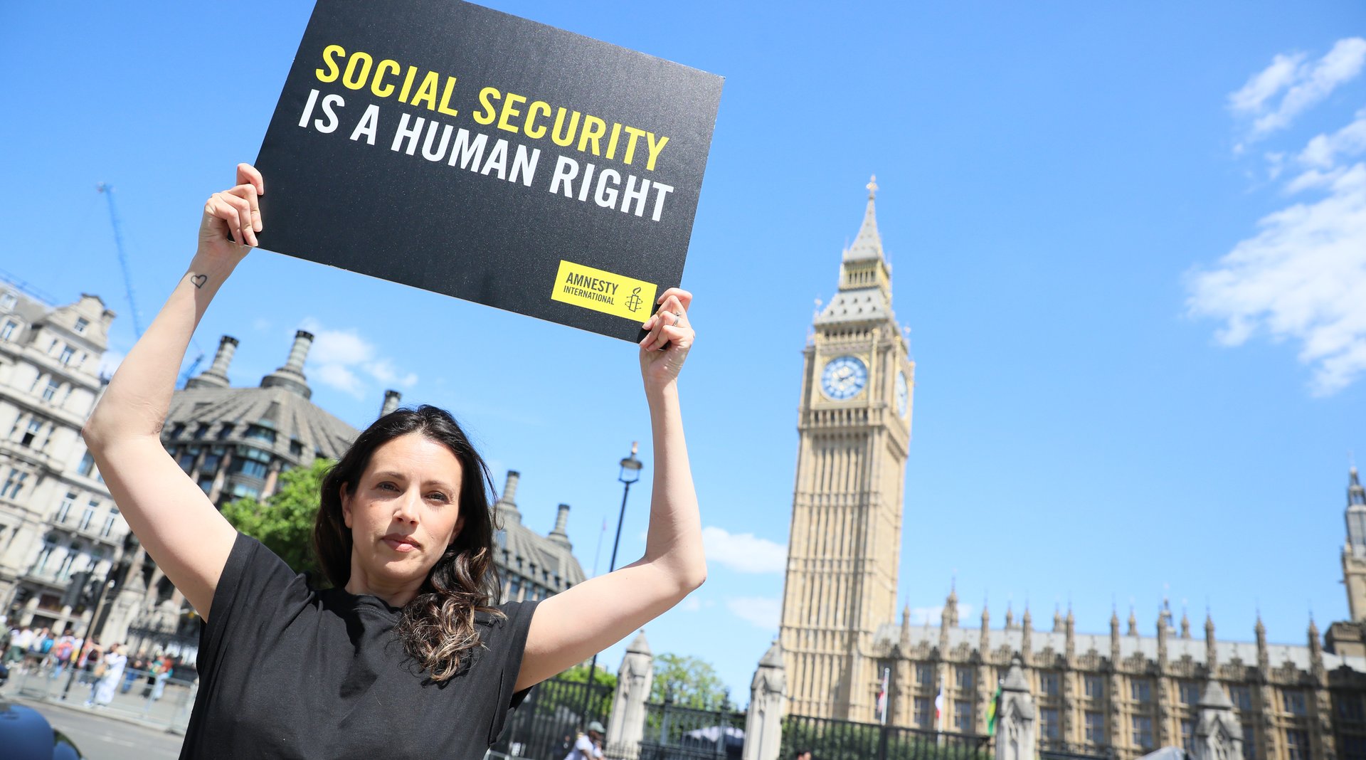 A woman in an Amnesty International UK t-shirt holds up a placard that says 'Social security is a human right' outside The Houses of Parliament.