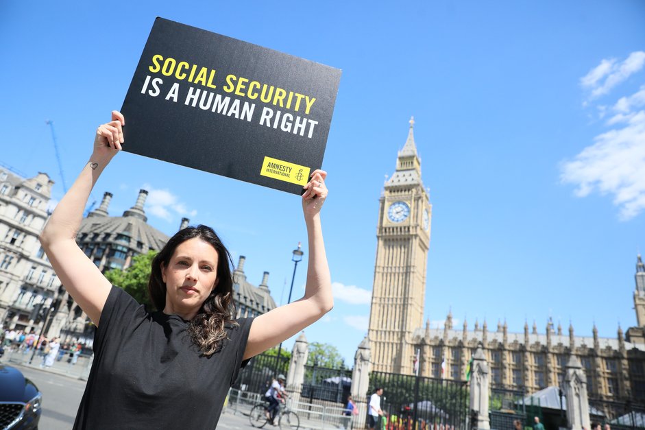 A woman in an Amnesty International UK t-shirt holds up a placard that says 'Social security is a human right' outside The Houses of Parliament.