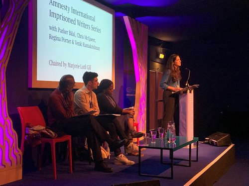 Image shows a woman reading at a lectern whilst four people sit on a stage