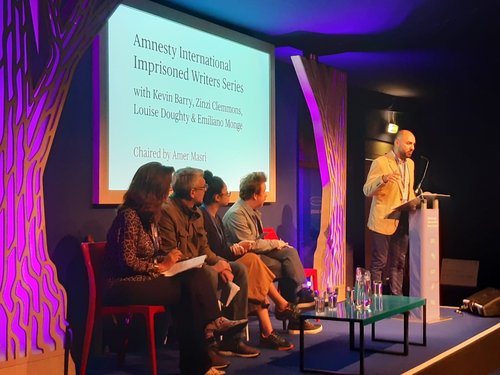 Image shows a man reading at a lectern whilst four people sit on a stage