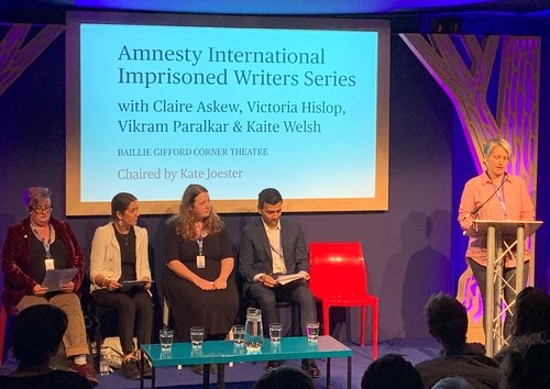 Image shows a woman reading at a lectern whilst four people sit on a stage