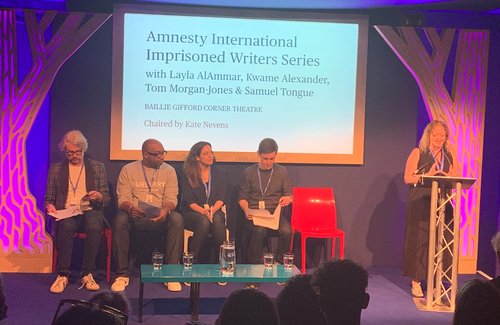 Image shows a woman reading at a lectern whilst four people sit on a stage