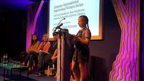 Image shows a girl reading at a lectern whilst four people sit on a stage