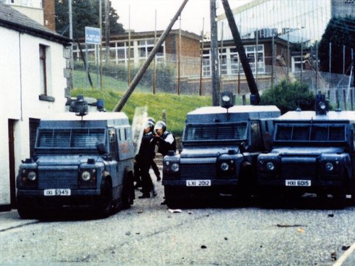 Three armoured paramilitary police vehicles parked on a street with three police officers wearing riot helmets and holding guns standing behind the vehicles