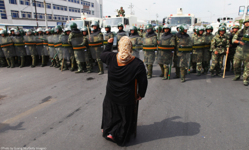 A person stands in front of a line of heavily armed soldiers who have their shields up. The person has their first in the air in defiance