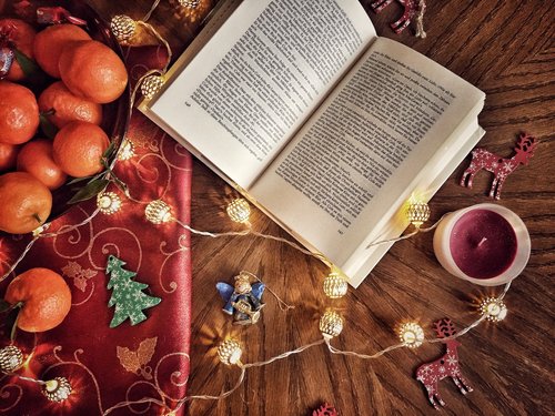 An open novel nestled next to a class of mulled wine, on a table with satsumas and gift wrapping trinkets.