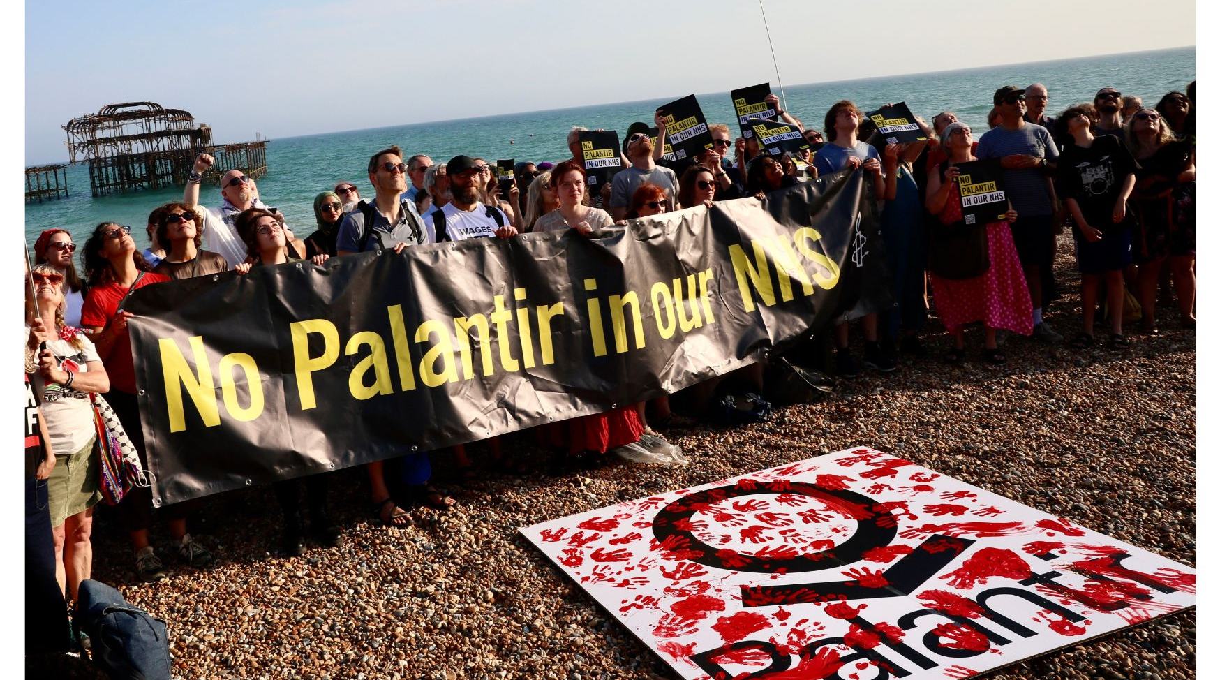 Crowd hold 'No Palantir in Our NHS' banner on Brighton beach