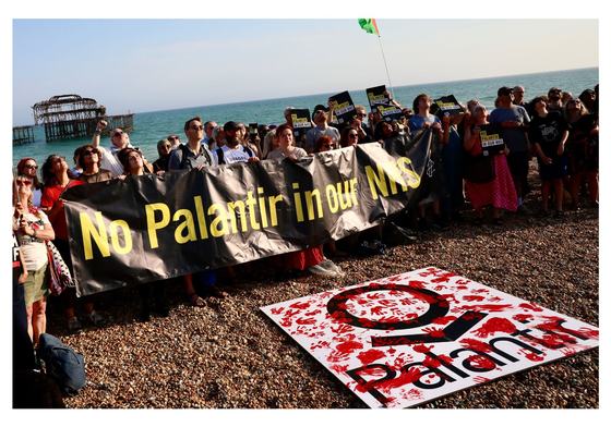 Crowd hold 'No Palantir in Our NHS' banner on Brighton beach