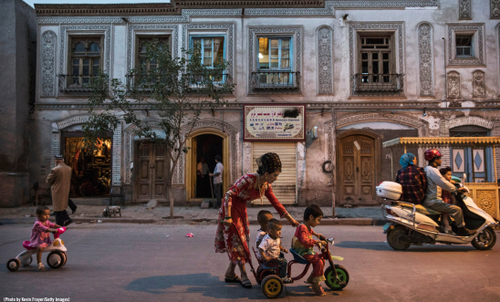 A street in North East China at nighttime shows a scene of a woman helping three children play on a small bike. Another chlld on a toy bike is behind them. The buildings are partially lit by what looks like a street light out of shot. There is also a person on a moped passing by the children