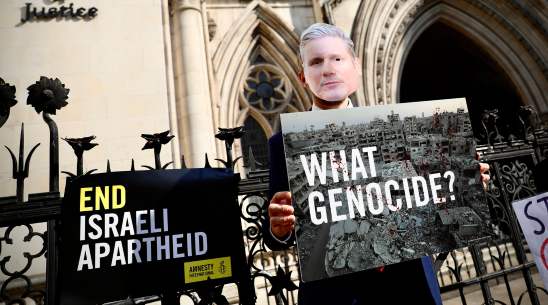 Person in Starmer mask holding placard questioning 'What Genocide?'