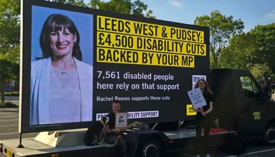 Image of an activist in front of a banner reading "LEEDS WEST & PUDSEY: £4,500 DISABILITY CUTS BACKED BY YOUR MP"
