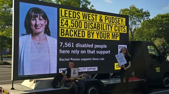Image of an activist in front of a banner reading "LEEDS WEST & PUDSEY: £4,500 DISABILITY CUTS BACKED BY YOUR MP"