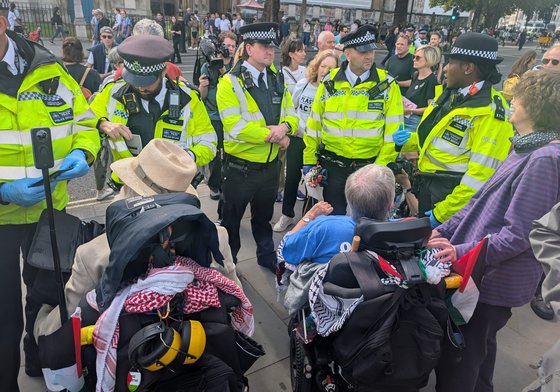 Circle of police people surround 2 people sitting on mobility scooters to arrest them