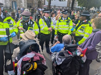 Circle of police people surround 2 people sitting on mobility scooters to arrest them
