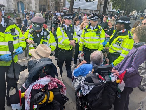 Circle of police people surround 2 people sitting on mobility scooters to arrest them