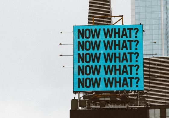 A large blue sign on top of a tall building reading 'what now?'