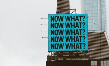 A large blue sign on top of a tall building reading 'what now?'