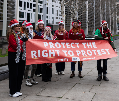 Carol singers wearing festive clothing hold a large red banner saying 'protect the right to protest' outside the Home Office